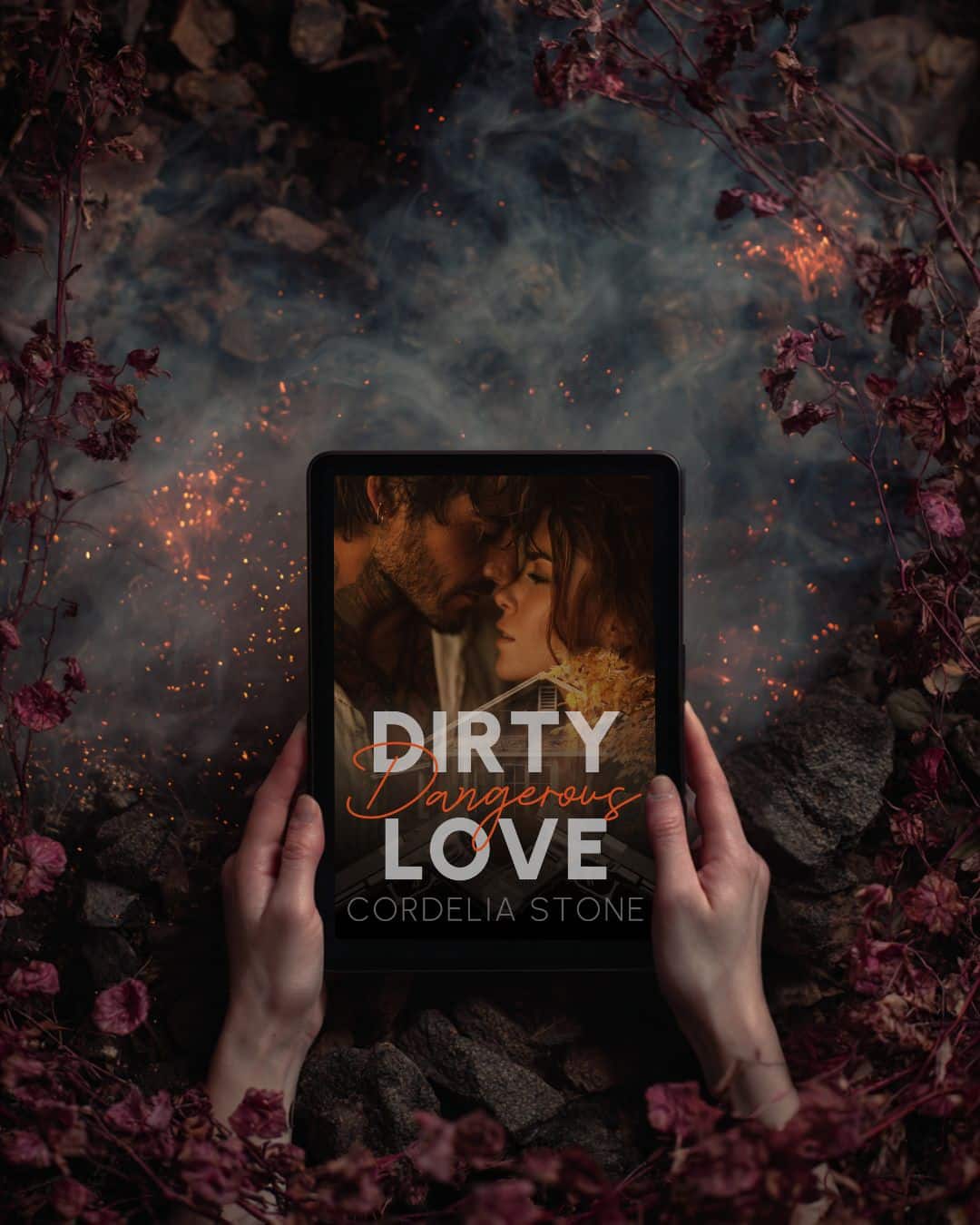 Olive Spencer, romance author - A person holds a tablet displaying a book cover titled “Dirty Dangerous Love” by Cordelia Stone, featuring a close-up of a man and woman about to kiss, surrounded by rocks, smoke, and red leaves.